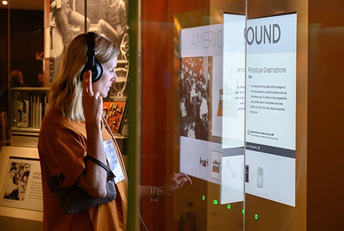 A woman with blond shoulder length hair wears headphones and stands in front of an interactive display about American Sound at the National Museum of American History.
