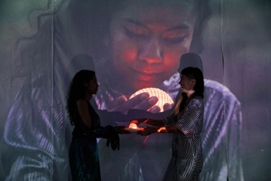 Two women stand facing each other. The woman on the right holds an illuminated round object. A screen in the background projects an image of a woman with wavy hair looking at an illuminated, striped, round object she holds in her hands.