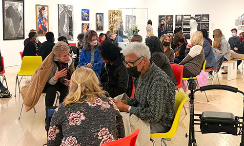 a group of people sit in yellow and orange plastic chairs in a museum with art hanging on the walls