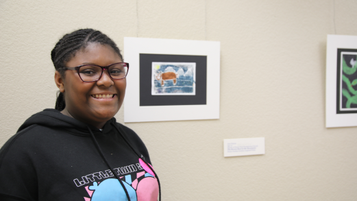 An African American woman with glasses and wearing a black hoodie stands in front of a small painting hanging on a beige wall