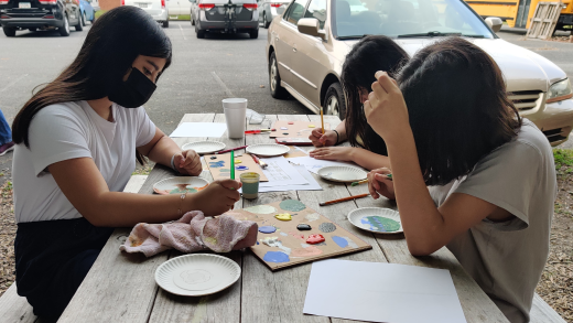 Three children sit at a wooden picnic table with paint supplies in front of them.