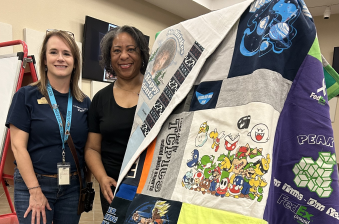 A white woman and an African American woman stand next to a quilt